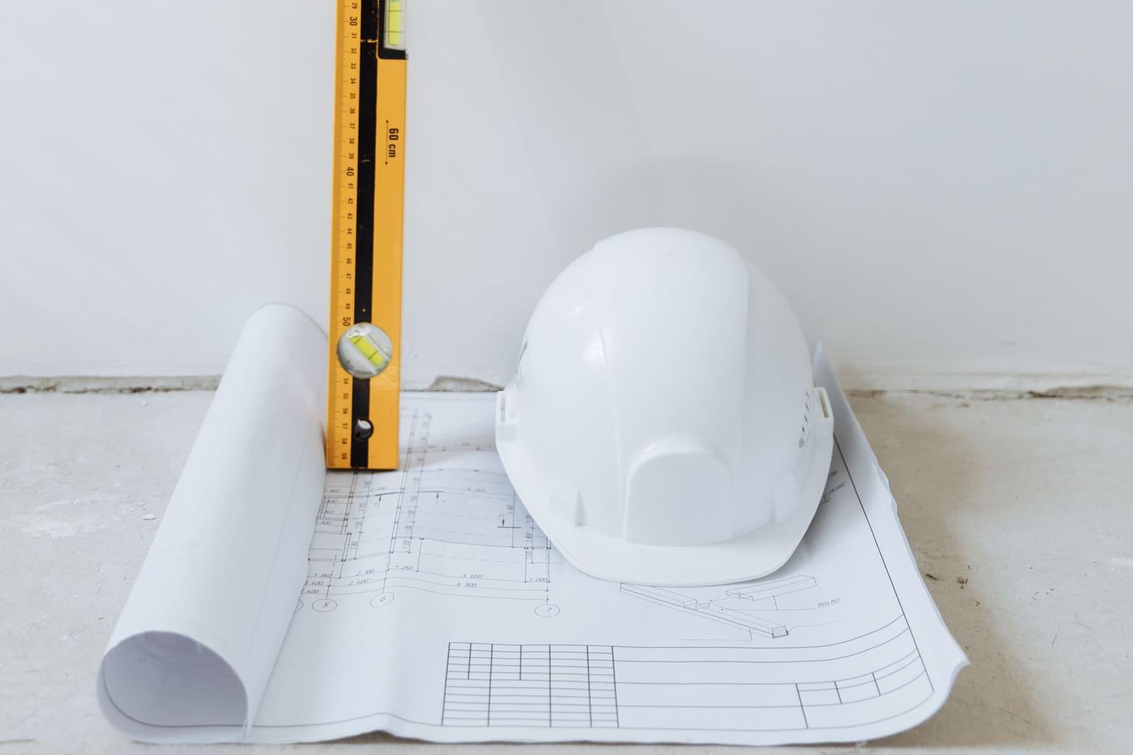 A white hard hat resting on rolled building plans next to an upright yellow spirit level on a plain concrete floor