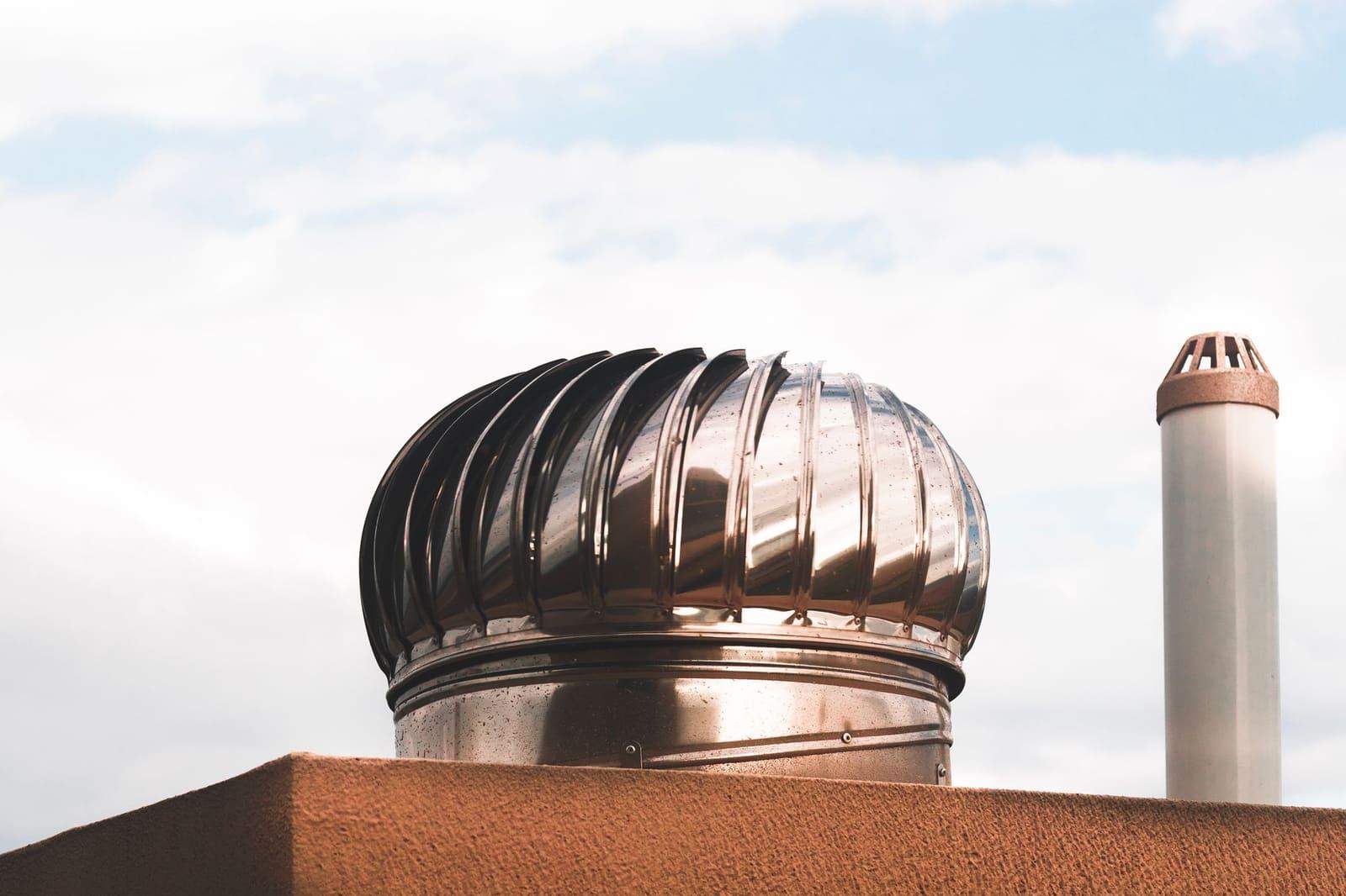 A shiny stainless steel turbine vent on a flat stucco rooftop beside a white vent pipe, under a bright hazy sky