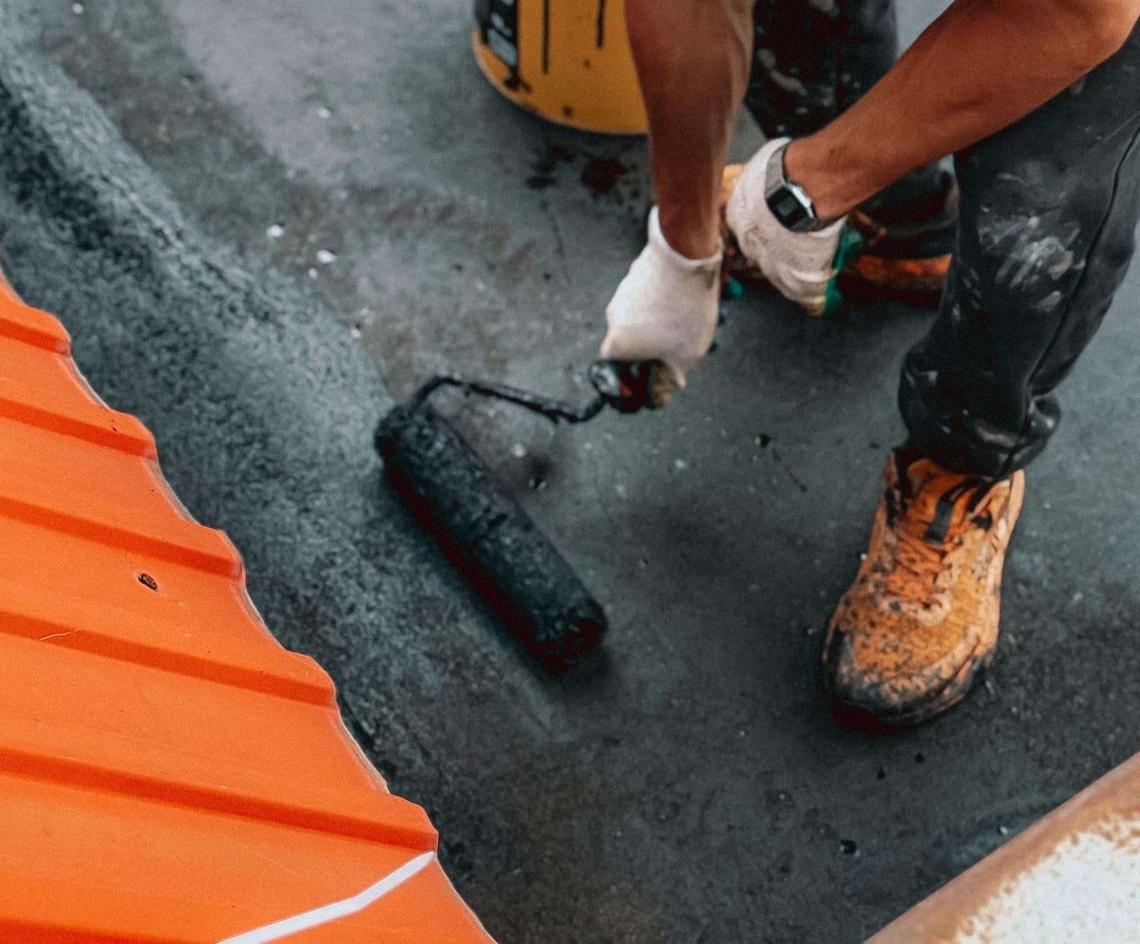 A gloved hand pulling a paint roller loaded with dark coating across a flat roof next to an orange metal edge