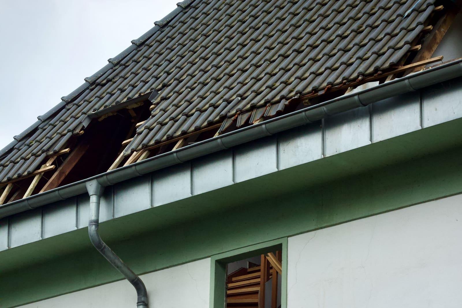 A gray tile roof with a ragged hole where tiles and sheathing have torn away, above a metal gutter and a white wall