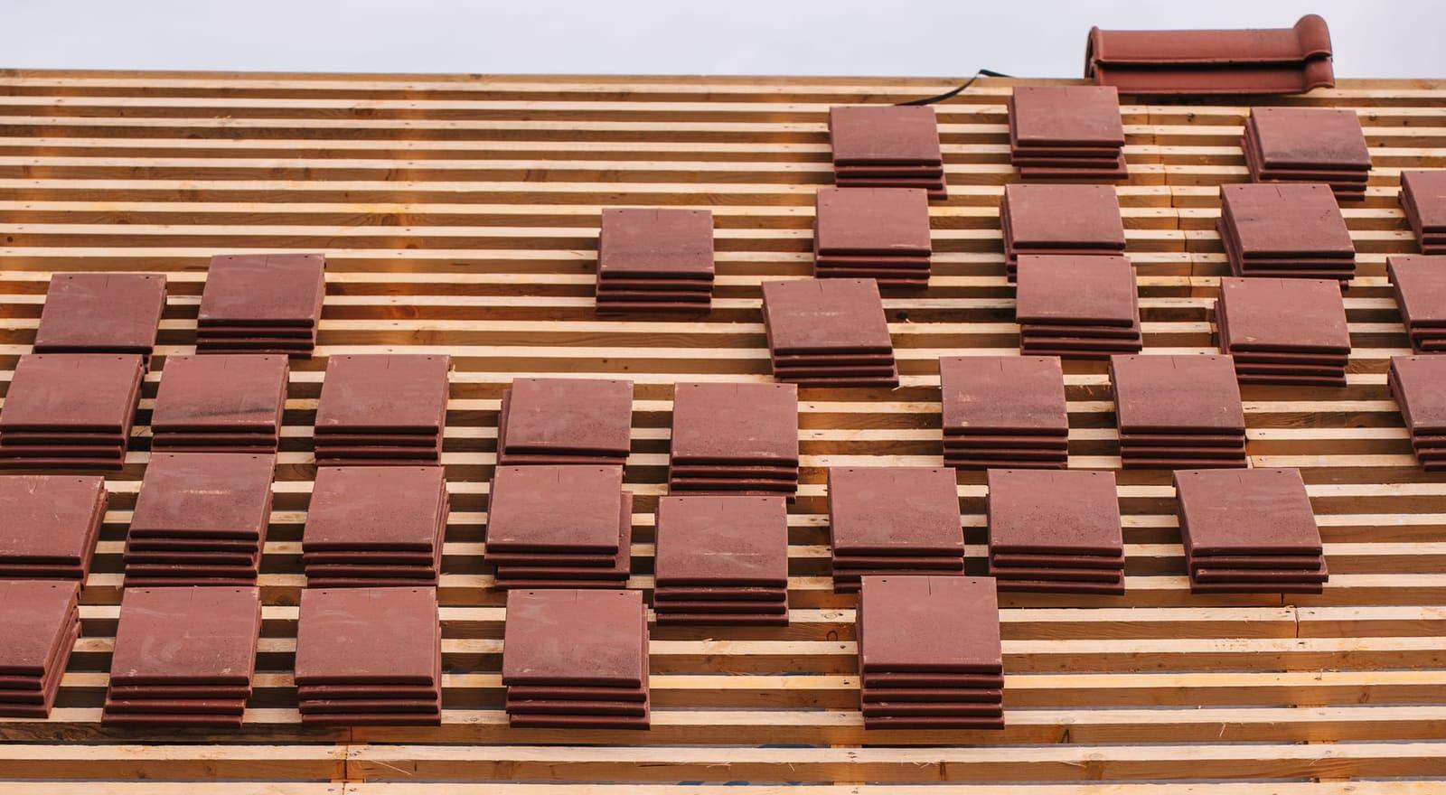 Stacks of red clay roof tiles set out on the wooden battens of a roof deck, with a ridge tile at the top