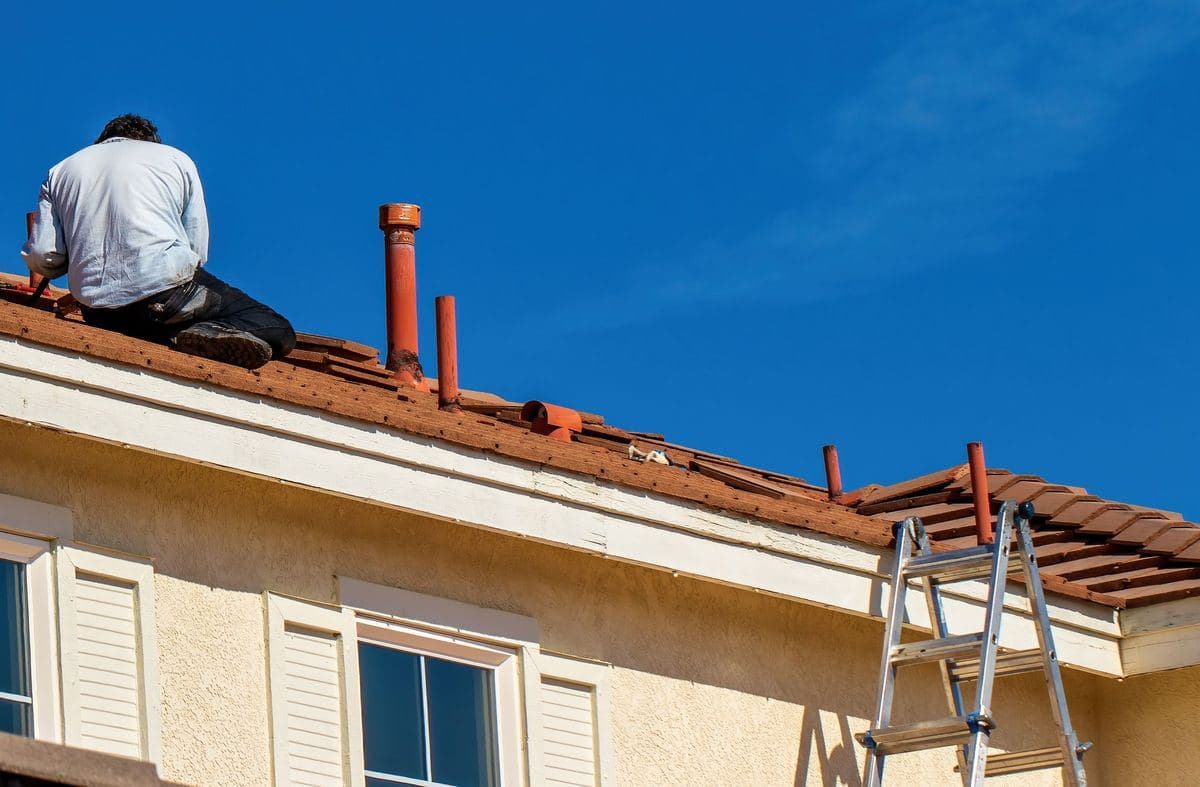 A roofer kneels on the clay tile roof of a two-story stucco house, beside vent pipes and loose tiles, with a ladder against the eave